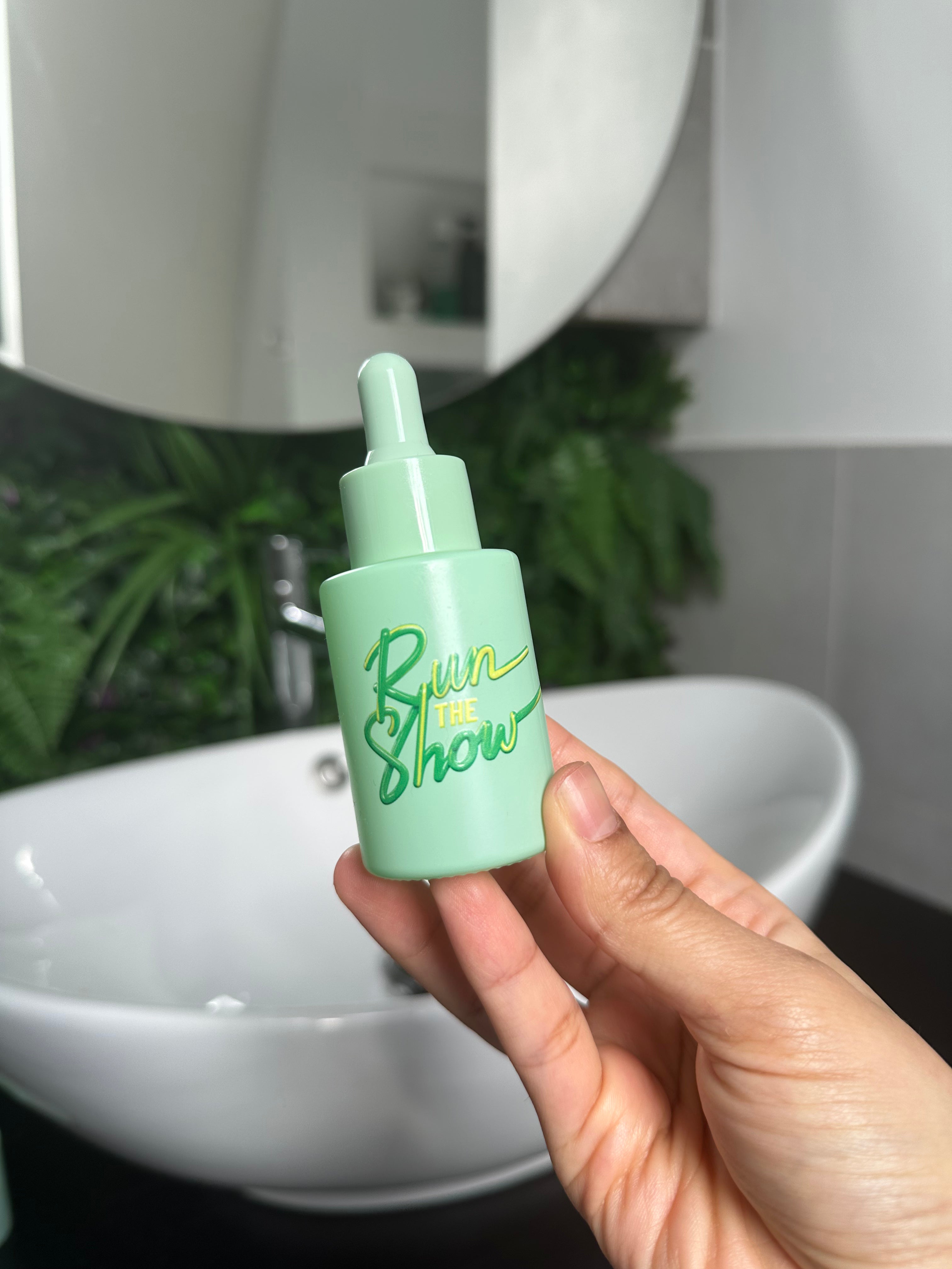 Person holding Run The Show Facial Oil in a bathroom setting – green dropper bottle captured in natural light, evoking focus and clarity through its calming scent.