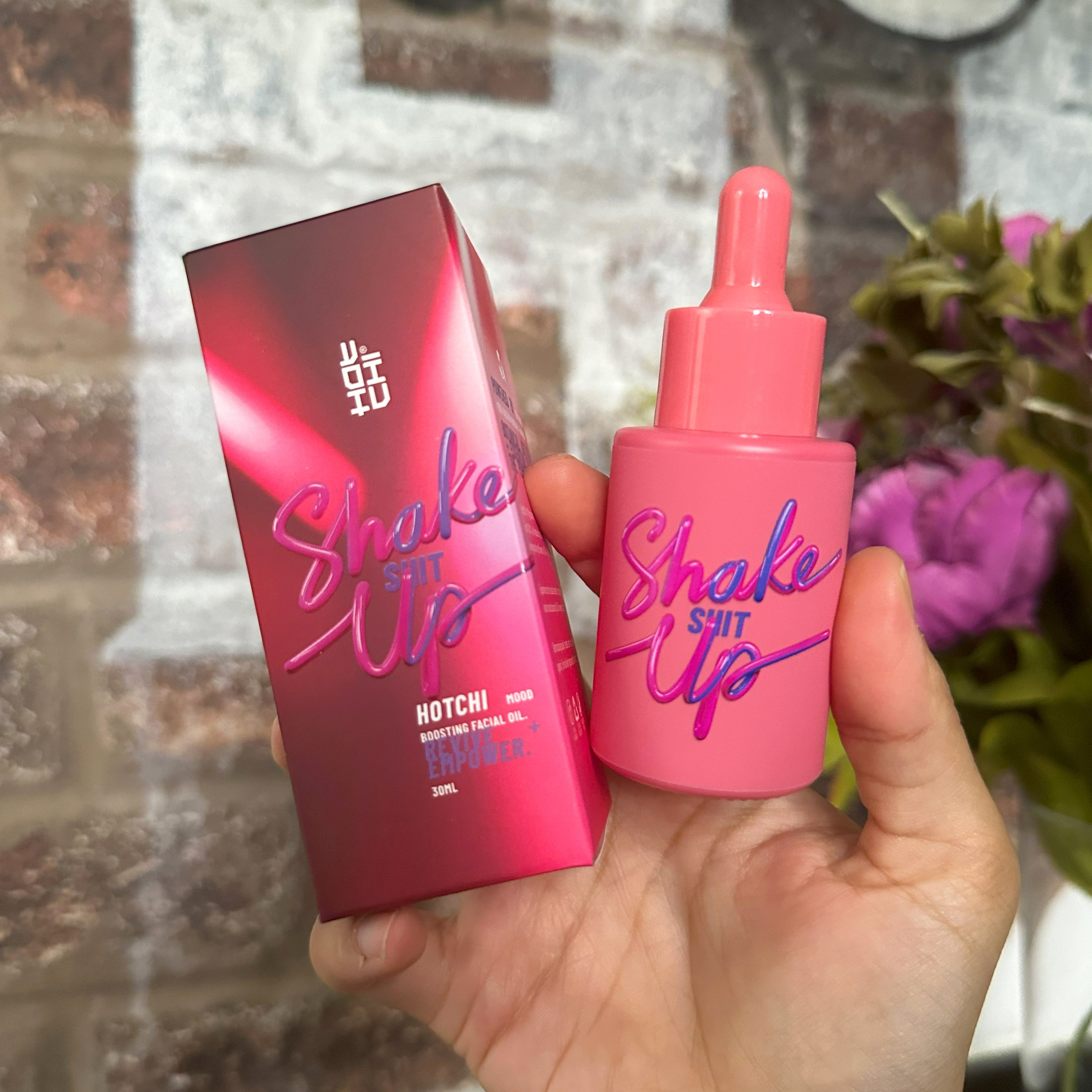 A hand holding the &#39;Shake Sh*t Up&#39; facial oil by HotChi, showing both the bright pink dropper bottle and its matching bold, vibrant packaging against a blurred background with flowers and a brick wall
