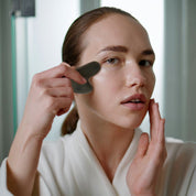 White woman gliding a stainless steel gua sha along her jawline, emphasizing facial contouring and tension relief.